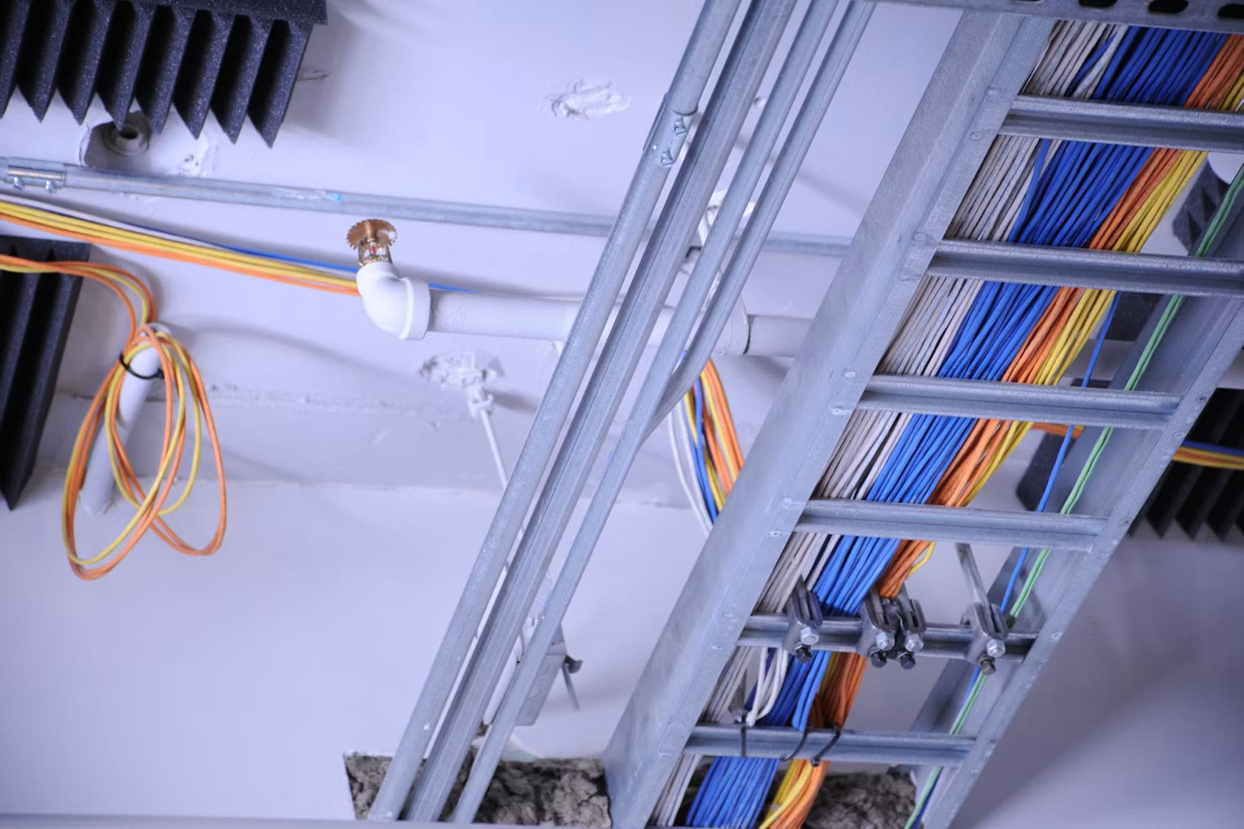 Kabelladder tegen een plafond, gevuld met gebundelde blauwe, grijze en oranje leidingen, met daarnaast stalen buizen en een sprinklerkop.