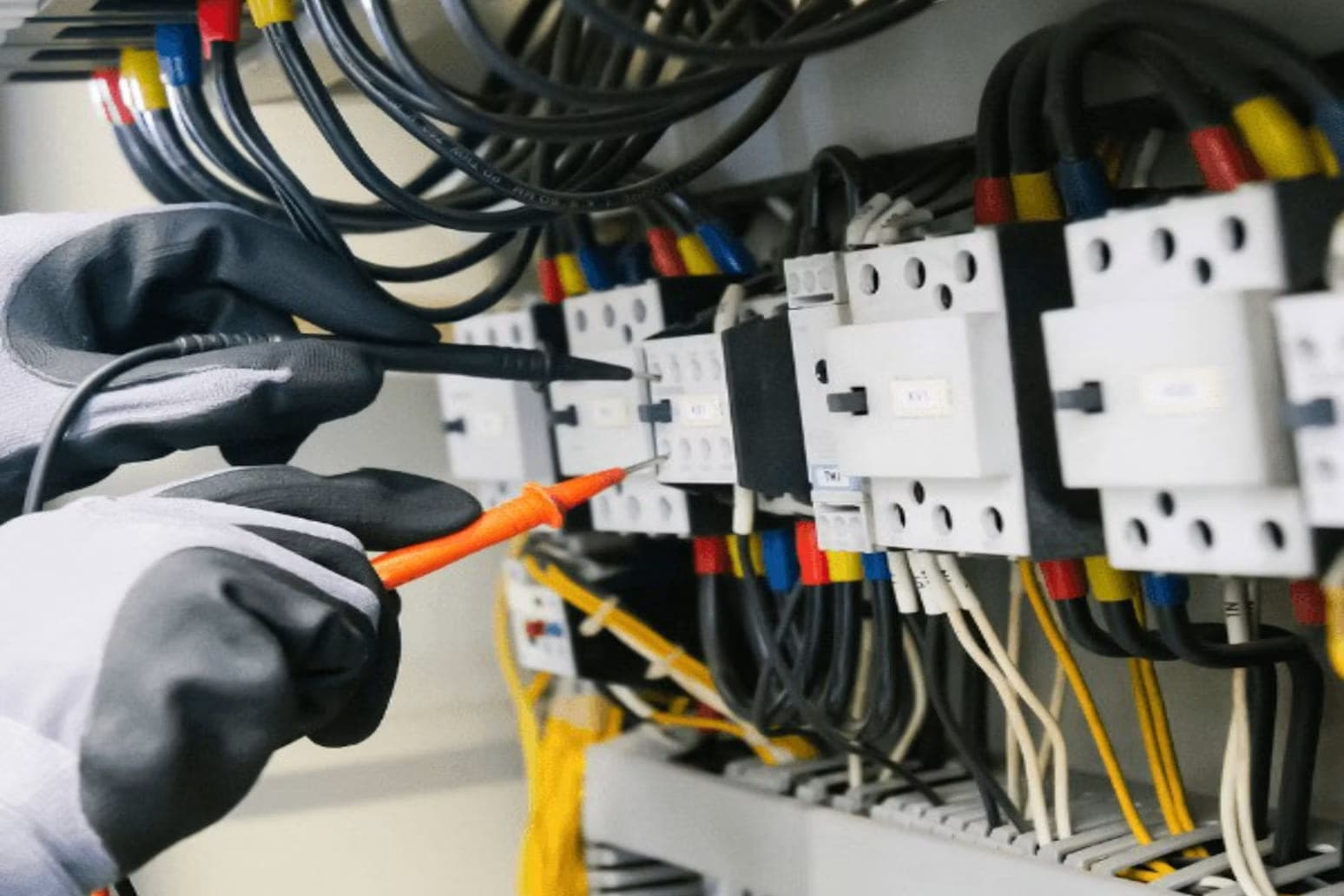 Meten in een schakelkast: met werkhandschoenen worden een zwarte en een oranje meetpen tegen de aansluitpunten van een rij magneetschakelaars gehouden.