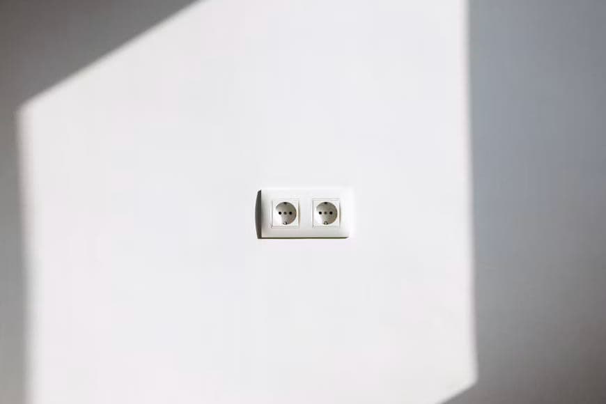 Dubbele wandcontactdoos op een witte muur, met een schuine baan zonlicht eroverheen.